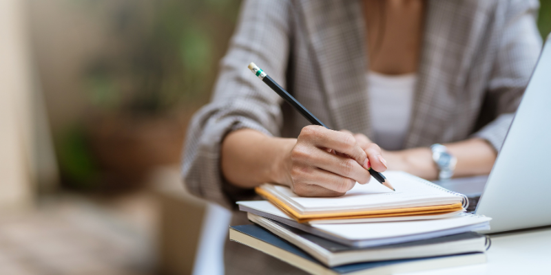 A close-up of a person writing in a notebook with a pencil, in front of a computer.