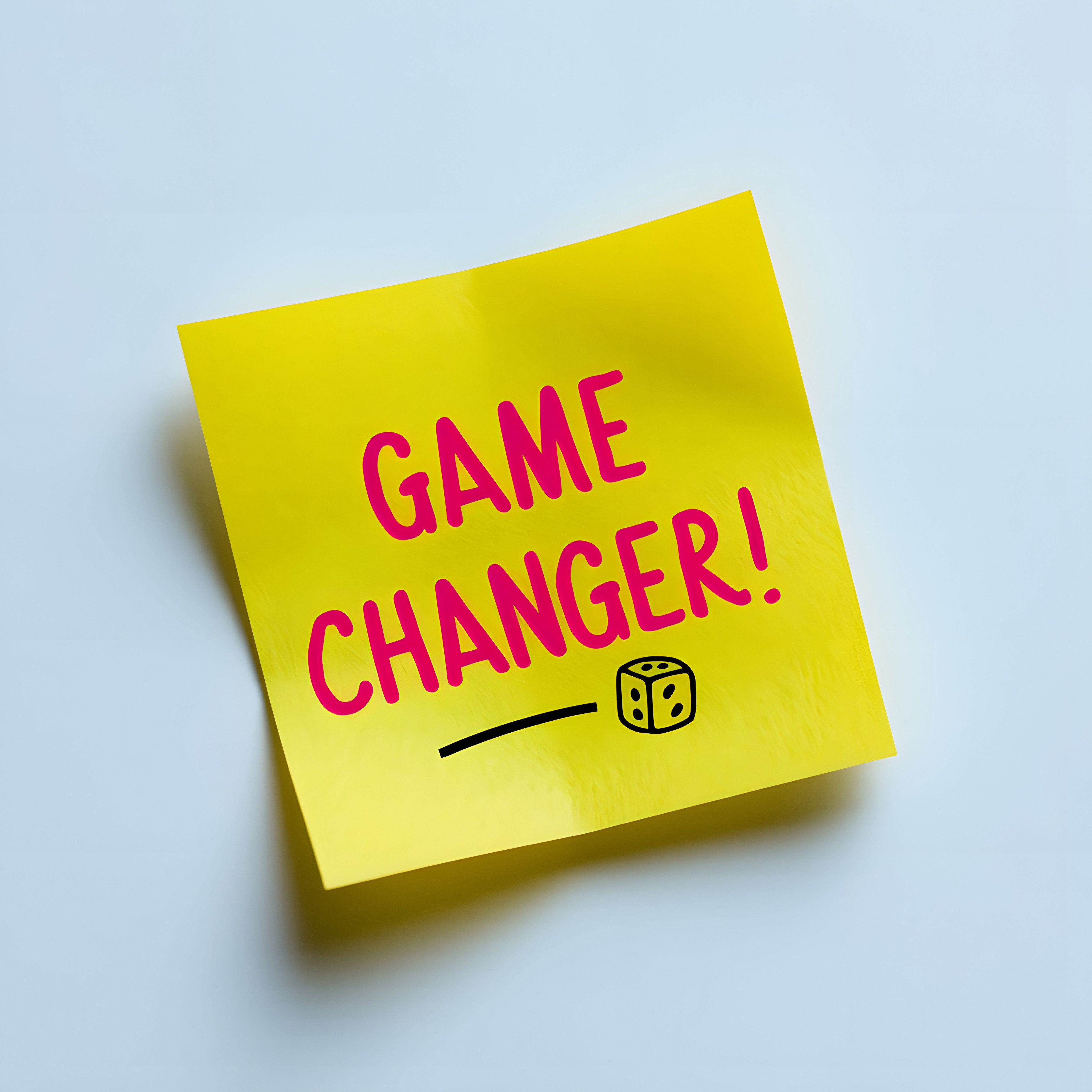 A yellow sticky note with the words 'GAME CHANGER!' written in bold pink letters. Below the text, there is a small illustration of a rolling die. The note is slightly curled and placed on a light blue background.